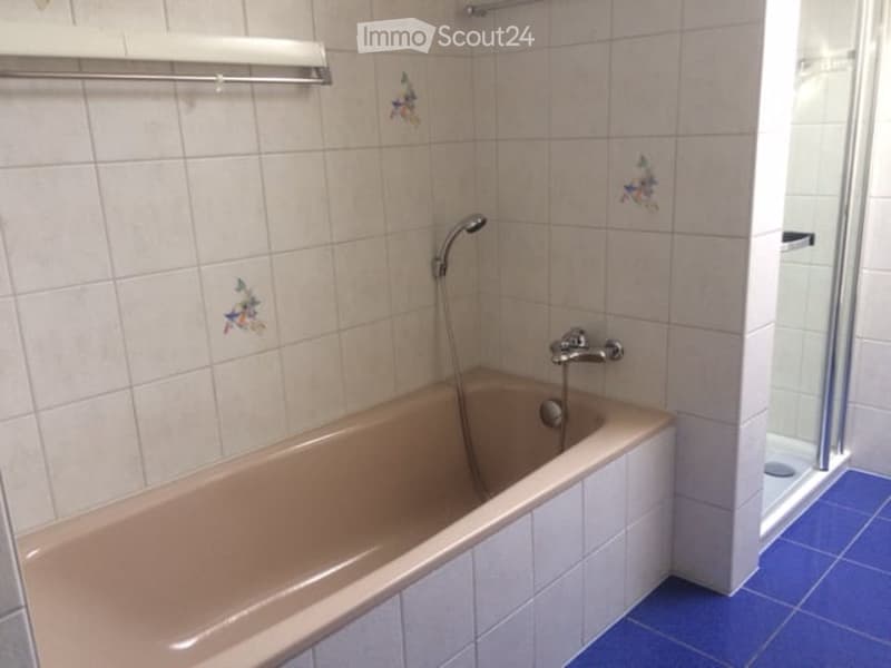 Bathroom with a beige bathtub, white tiled walls, and a blue tiled floor. A showerhead is attached to the wall above the tub, and a small decorative tile design is visible on the wall.