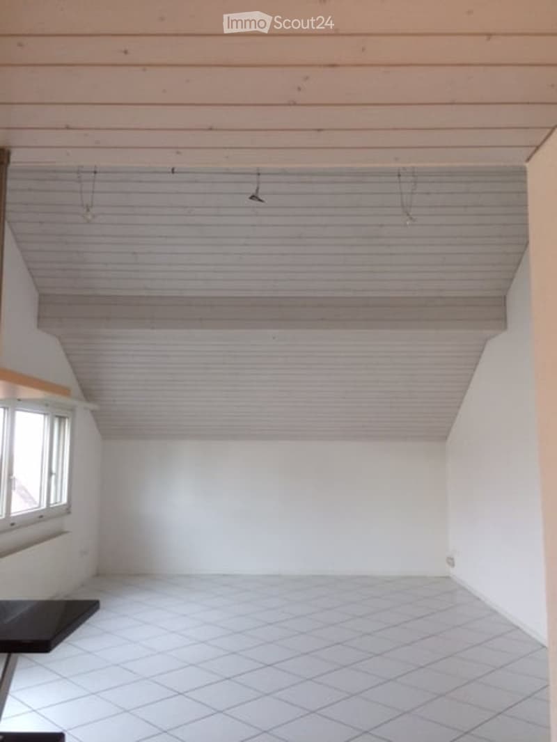 Empty room with white tiled floor, white walls, and a window on one side. The ceiling has exposed wooden beams and a slanted section.