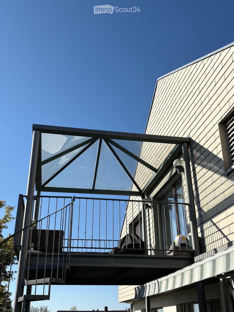 Balcony with a modern metal railing, glass roof structure, and outdoor furniture including a chair, table, and grill.