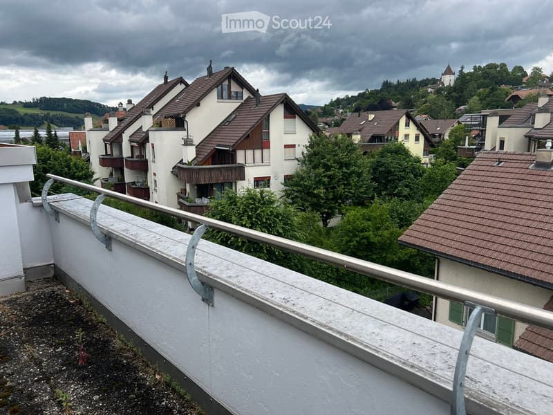 Balcony with metal railing overlooking a residential area with houses, trees, and a distant view of a town and hills.