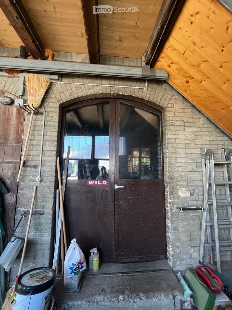 An unfurnished room with a wooden ceiling and exposed beams, featuring a large wooden door with glass panels, a broom, and various tools and items scattered around.
