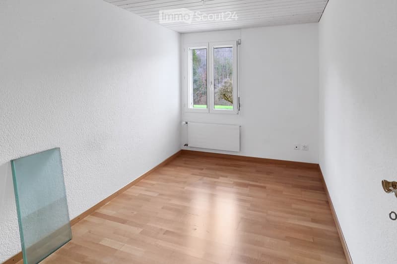 Empty room with light wooden flooring, white walls, and a large window with a view of greenery. A radiator is mounted on the wall below the window, and a glass partition is visible on the left side.