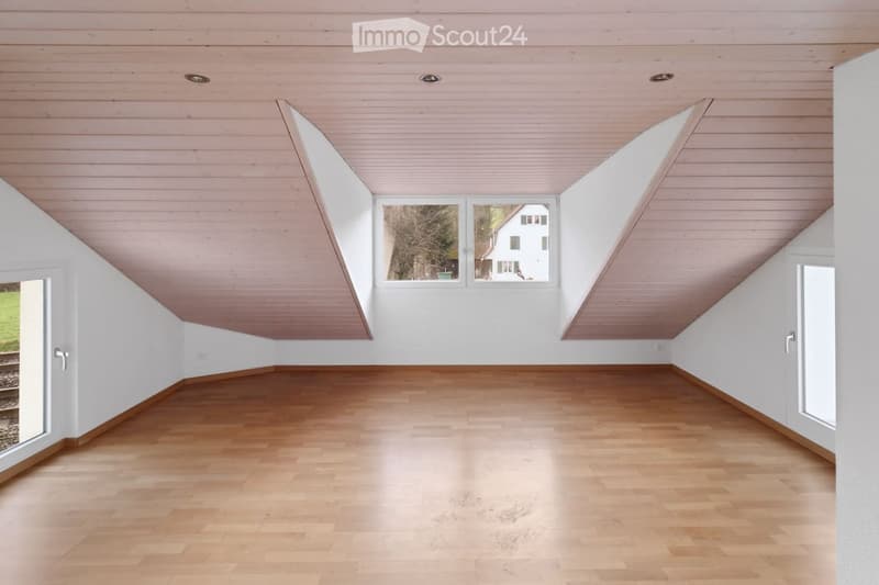 Empty room with wooden flooring and white walls, featuring a slanted wooden ceiling and large windows offering a view of the outside.