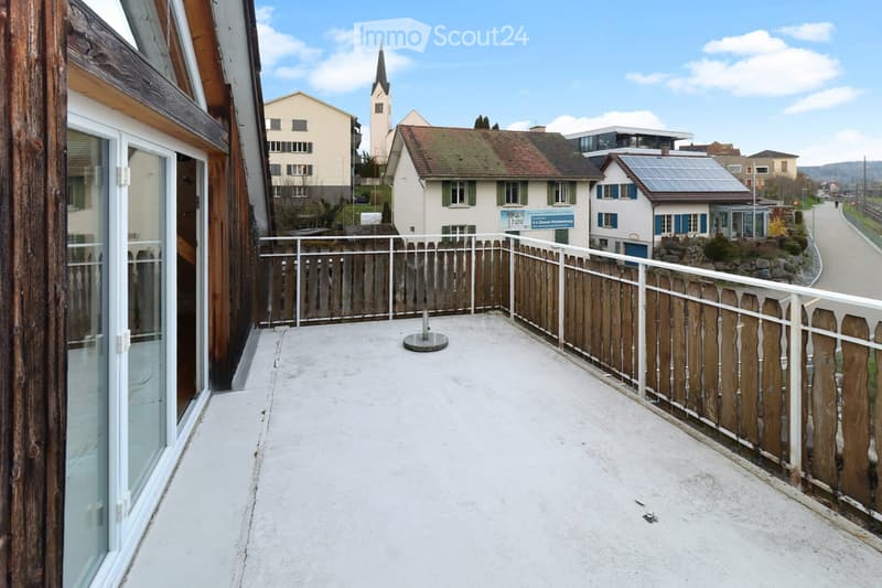 Balcony with wooden and metal railings, concrete floor, overlooking a residential area with houses and a church tower.