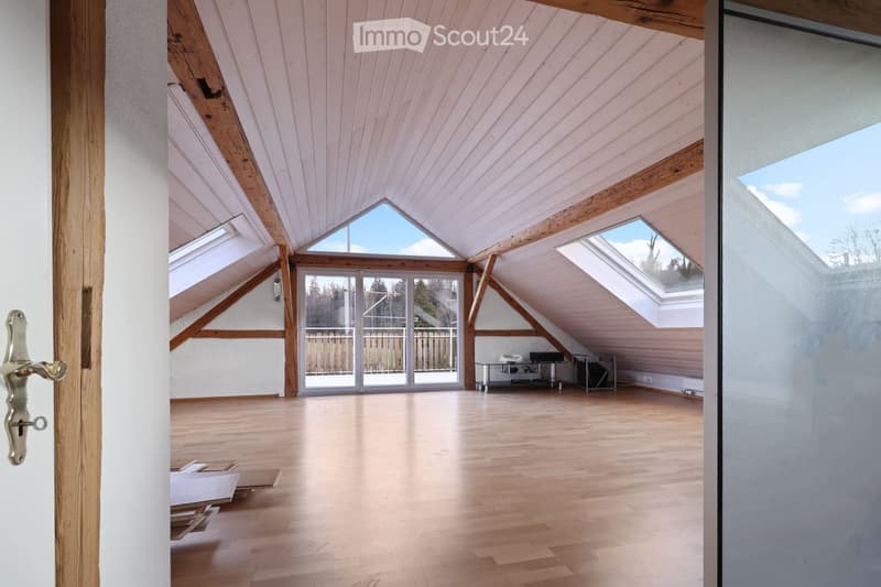Bright, spacious attic room with wooden beams, skylights, and large windows, featuring a polished wooden floor and white walls.
