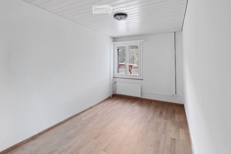 Empty room with light wooden flooring, white walls, and a single window with a view of an outdoor area. The room has a white radiator and a ceiling light fixture.