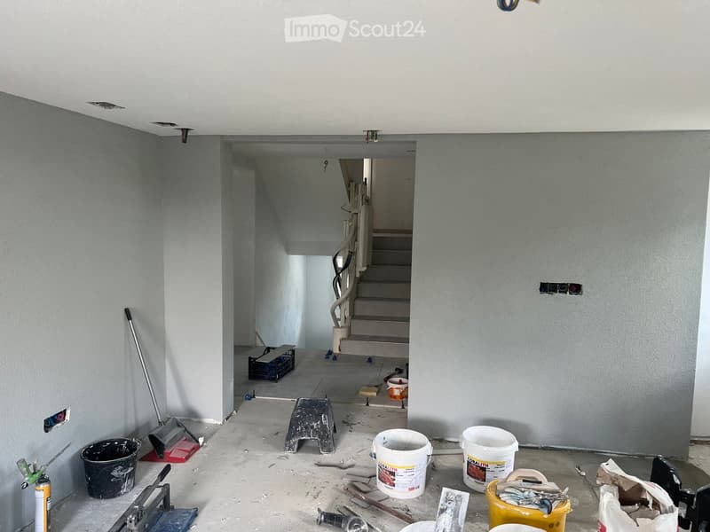 Empty hallway with unfinished walls and ceiling, visible staircase leading upwards, construction materials and tools scattered on the floor.
