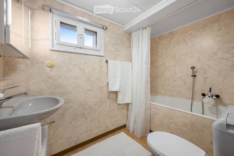 Bathroom with a sink, bathtub, toilet, and white towels. Features a window with a curtain and a small white rug on the floor.