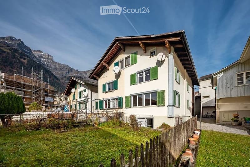 A two-story house with white walls, green shutters, and a wooden roof, situated in a scenic mountain village with a grassy front yard and wooden fence.