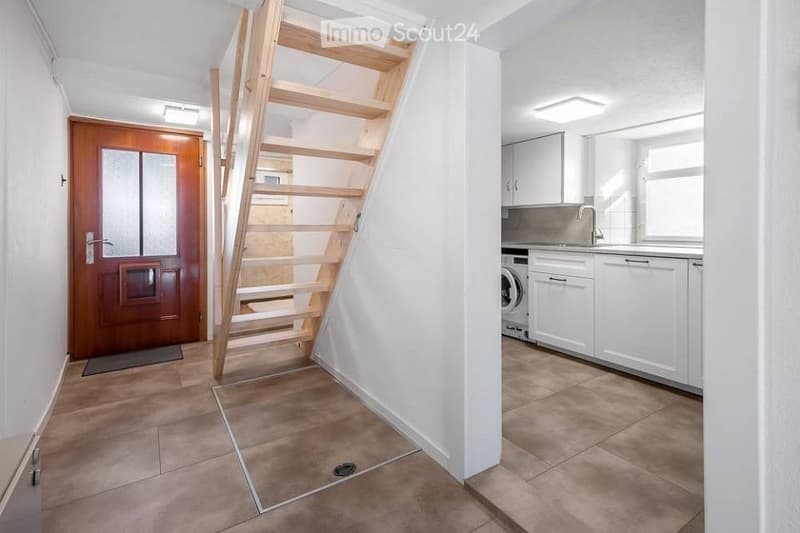 A narrow hallway with a wooden staircase leading upwards, a wooden front door, and a view into a modern kitchen area with white cabinets and a washing machine.