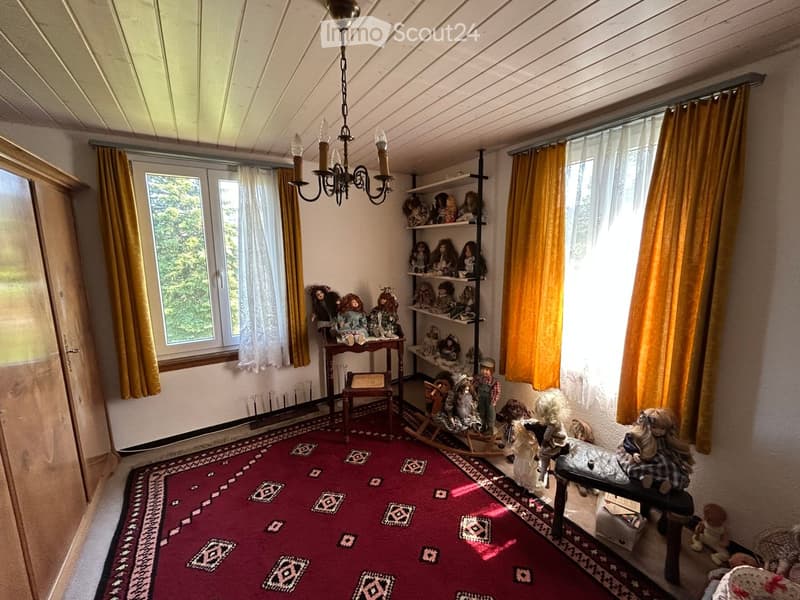 A cozy room with a red patterned rug, wooden furniture, shelves filled with dolls, and a chandelier hanging from the ceiling.