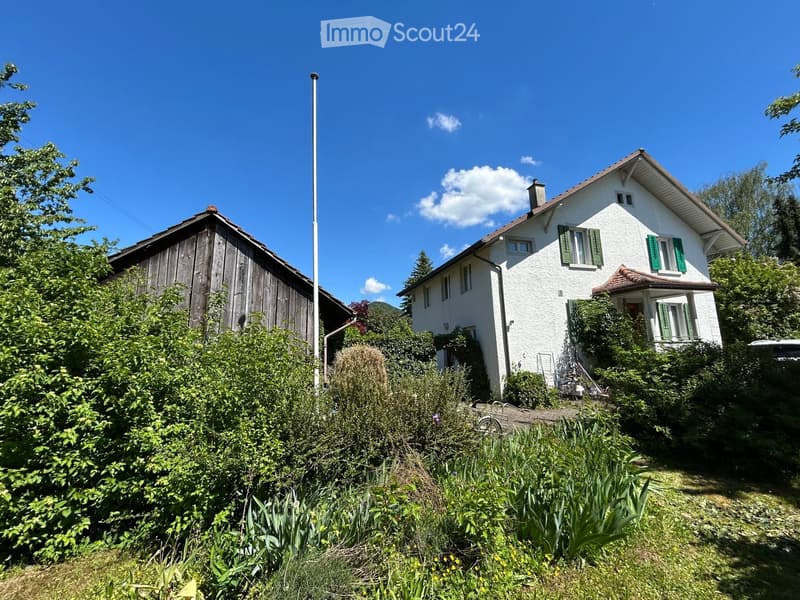 Two-story house with white exterior, green shutters, and a wooden outbuilding surrounded by lush greenery and a clear blue sky.