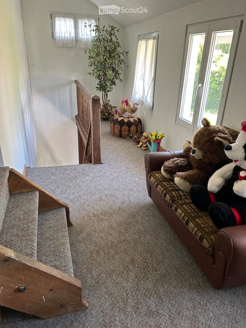 Carpeted living room with a brown sofa, large stuffed animals, a wooden staircase, and decorative items including a plant and a toy chicken.