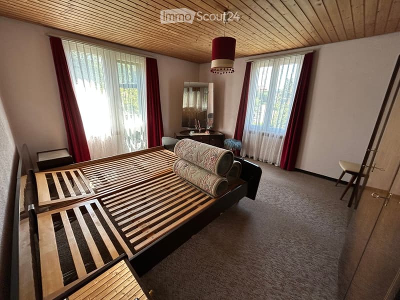 Bright bedroom with wooden bed frame, neatly stacked bedding, large windows with red curtains, a mirror, and a small table with decorative items.