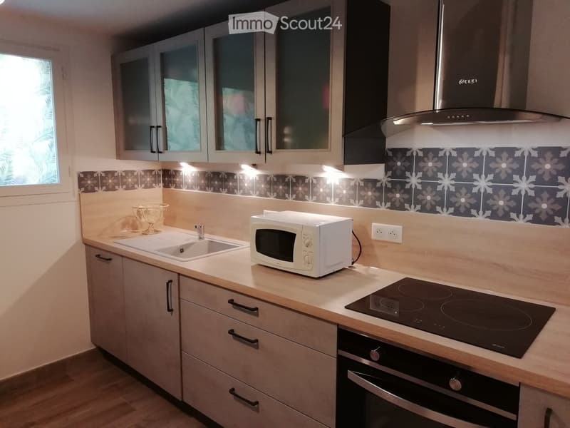 Modern kitchen with light wood cabinets, stainless steel appliances, and a decorative backsplash. Features include an induction stove, microwave, sink, and a bowl on the counter.