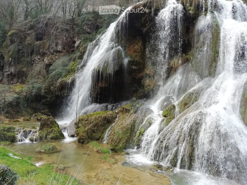 A natural waterfall cascading over moss-covered rocks, surrounded by greenery and a small stream below.