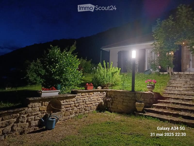 Nighttime garden with stone wall, potted plants, steps leading to a house, and outdoor lighting.