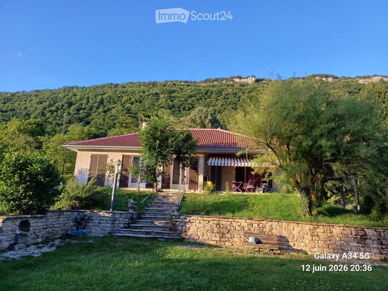 A two-story house with a red-tiled roof, surrounded by a well-maintained garden and lush greenery. The house features a covered porch area with outdoor furniture and is situated on a raised ground floor with stone steps leading up to it.