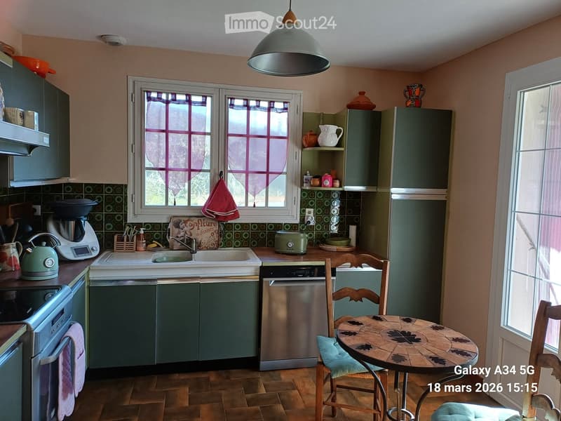 A cozy kitchen with green cabinets, a stainless steel dishwasher, a coffee maker, a toaster, a teapot, and a small dining area with a round table and chairs.