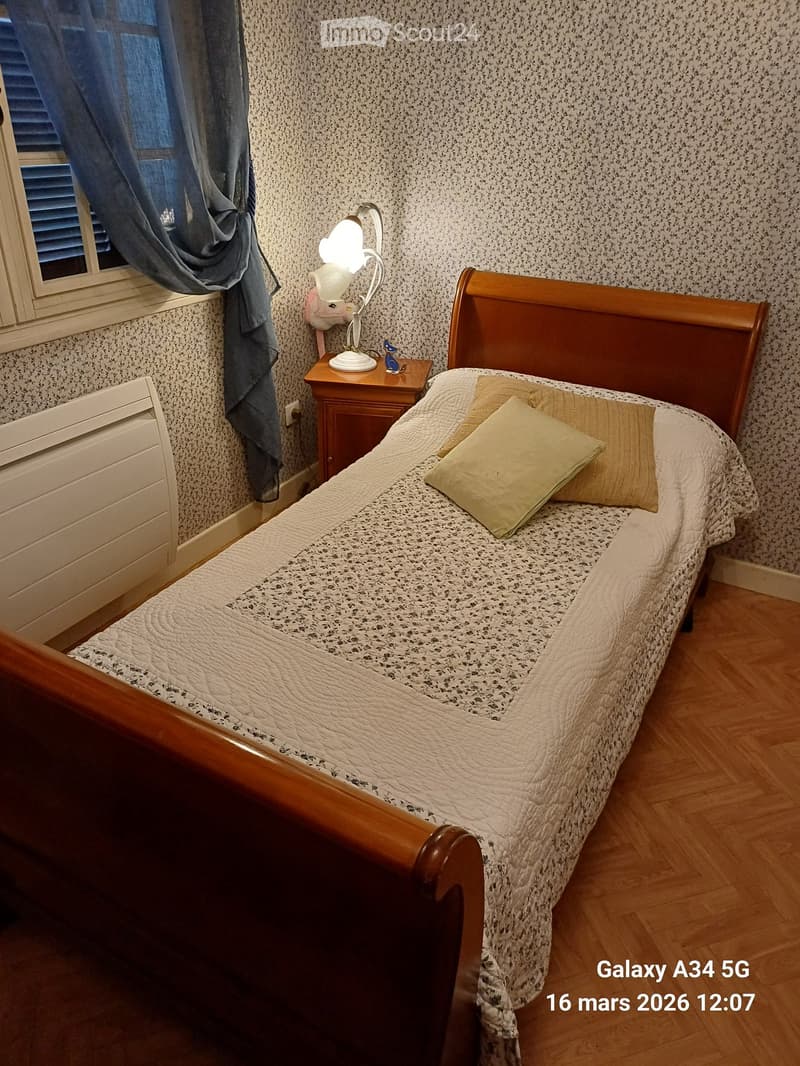 Cozy bedroom with a wooden bed, patterned bedding, nightstand, and lamp. The room features wooden flooring and patterned wallpaper.