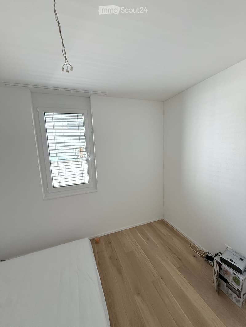 A furnished room with a wooden floor, white walls, a window with blinds, a bed, a printer, and a stack of books.