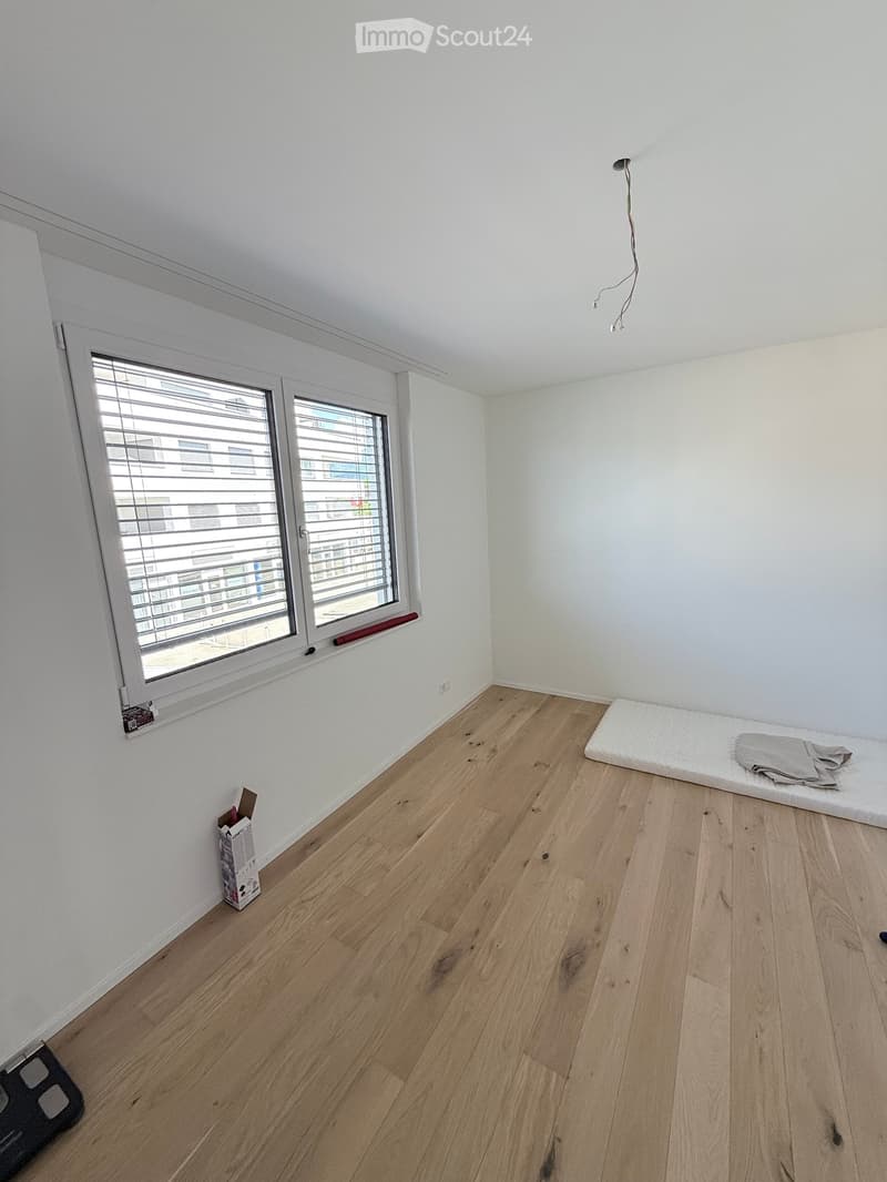 Empty room with wooden flooring, white walls, and large windows with blinds. A mattress and a folded blanket are placed in the corner, along with a small box on the floor.