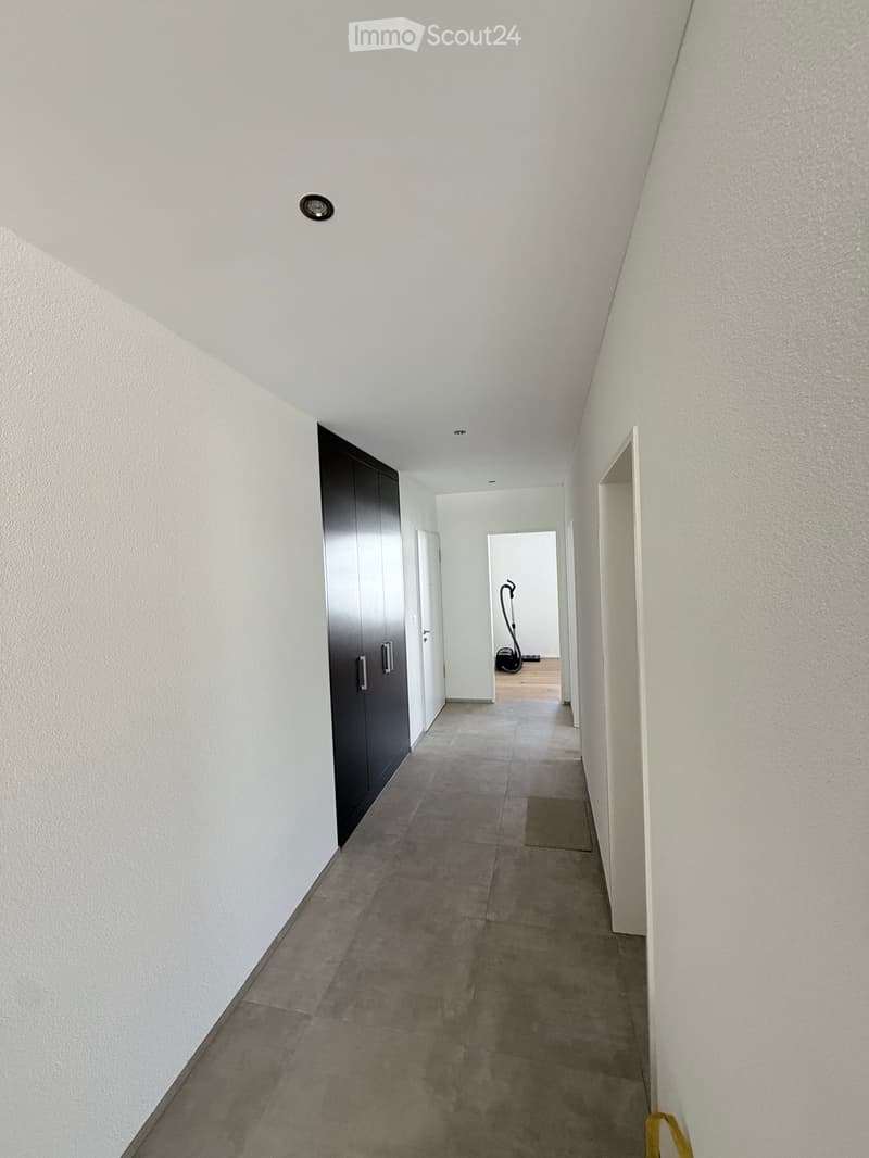 A clean, modern hallway with white walls, grey tiled floor, and recessed lighting. Features a dark wardrobe on the left and an open doorway leading to another room.