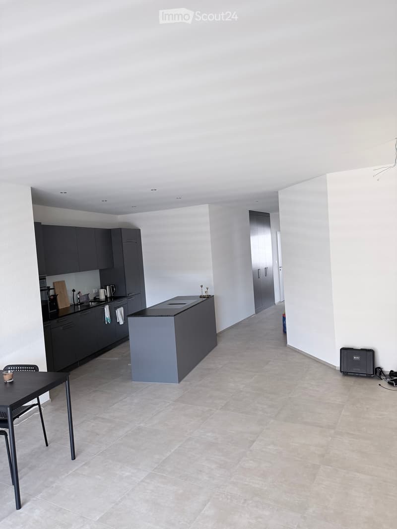 Modern kitchen with dark grey cabinets, a central island, a black countertop, and a small dining area with a table and chair. The floor is tiled, and the walls are white.
