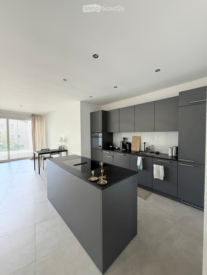 Modern kitchen with grey cabinets, black countertop, built-in oven, sink, and small dining area with table and chairs.