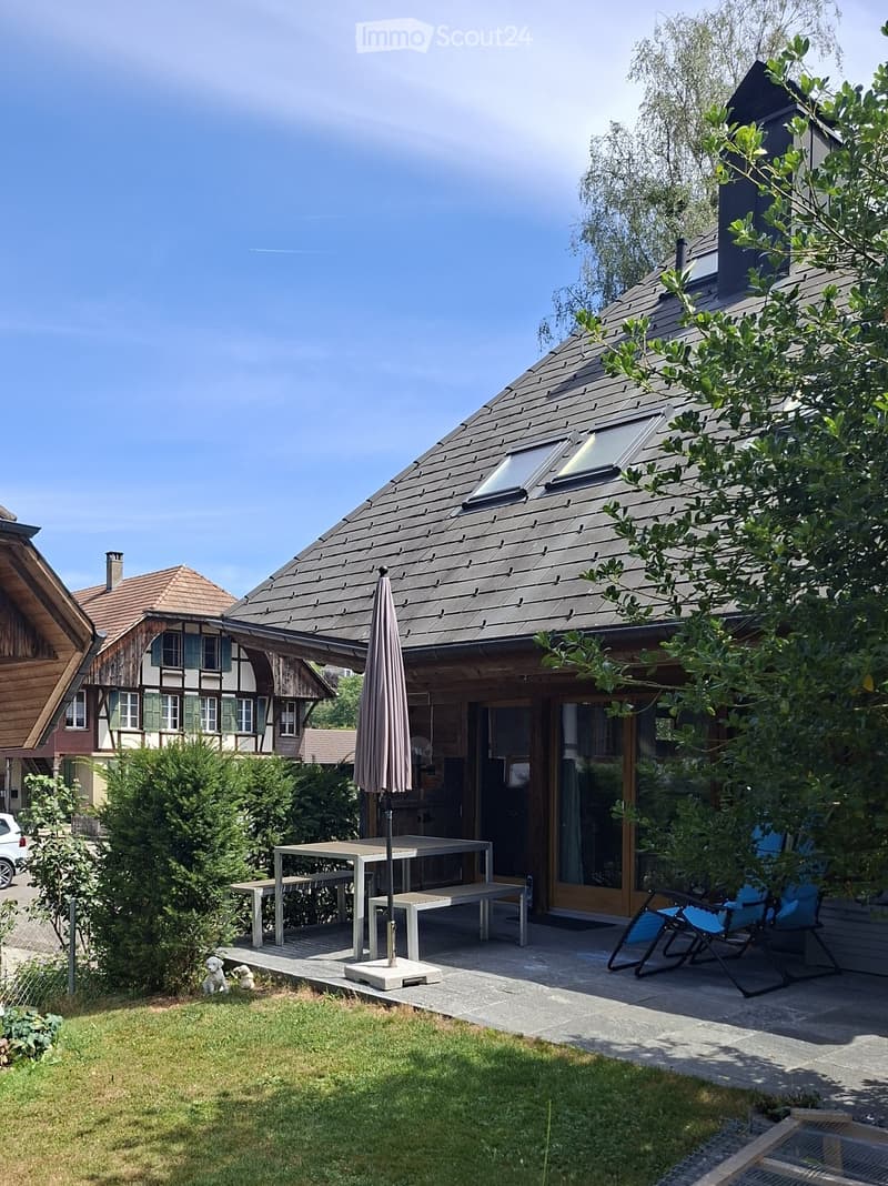 Outdoor patio area with a table, benches, chairs, and an umbrella, adjacent to a modern house with a sloped roof and large windows.