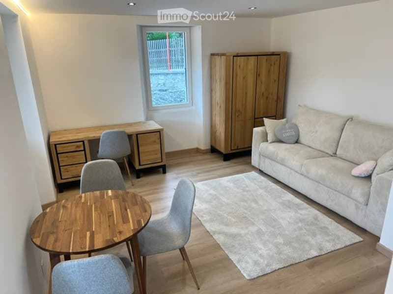 A modern, well-furnished room with a sofa, coffee table, chairs, a desk, a cabinet, and a small round table with chairs. The room features light wooden flooring and a neutral color palette.