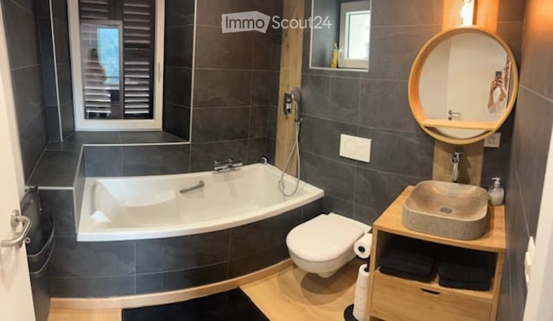 Modern bathroom with dark grey tiles, a bathtub, toilet, sink, and a round mirror above the sink. Wooden shelf holds towels and toiletries.