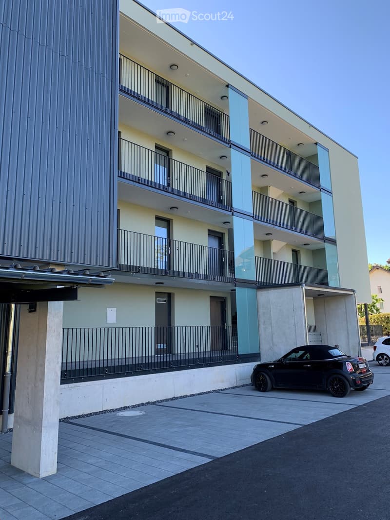 Modern multi-story apartment building with balconies, a covered parking area, and a small black convertible parked in the driveway.