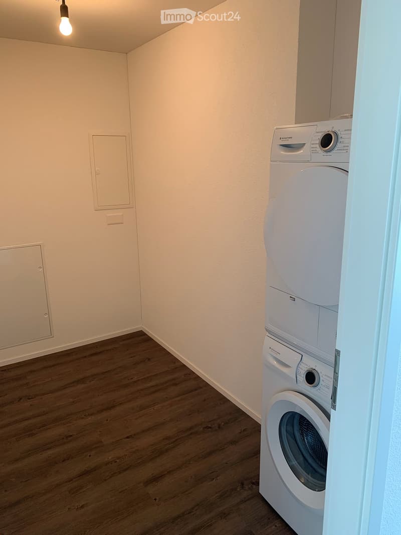 A small, well-lit room with wooden flooring and white walls, featuring a white washing machine and dryer unit.