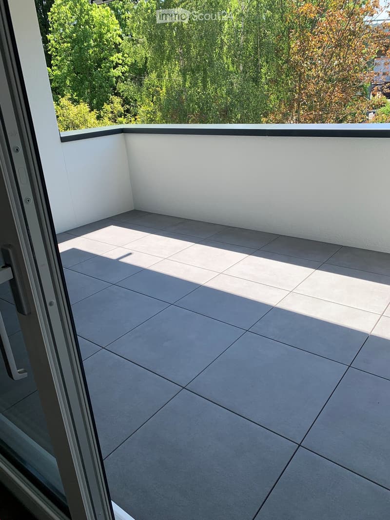 Modern balcony with grey tiled floor, white walls, and a black safety railing, overlooking a green, tree-filled area.
