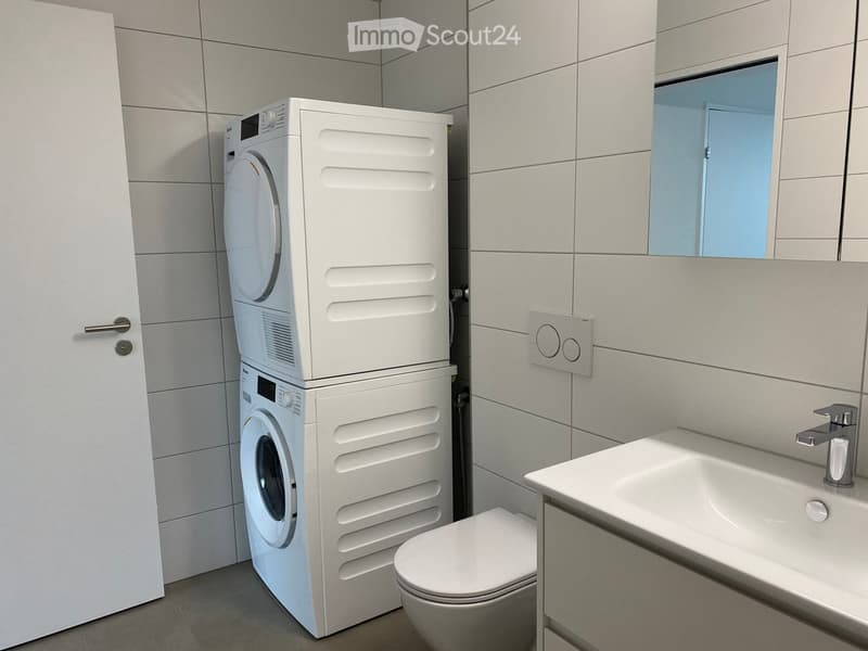 A small bathroom with white tiled walls, a white door, a white sink, a toilet, and a stacked washer and dryer.