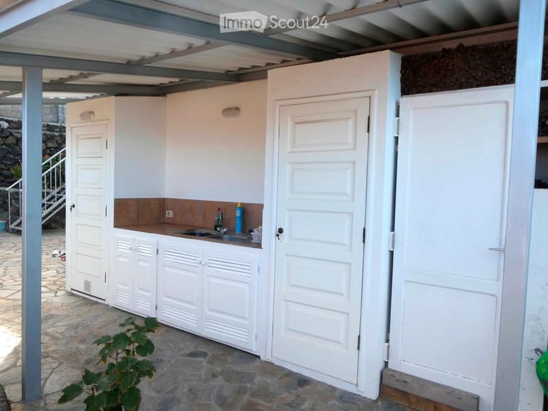 Compact outdoor kitchen with white cabinets, a sink, and a small countertop, featuring a stone floor and a metal roof structure.