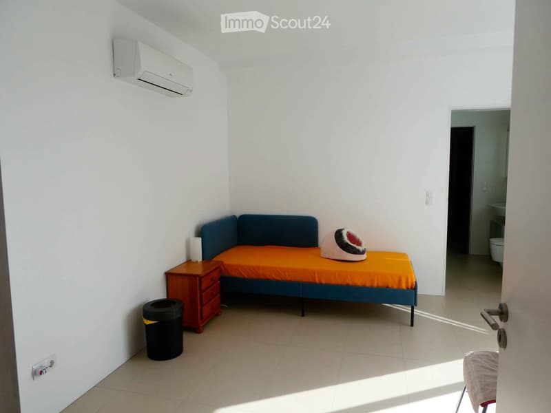 A small, furnished bedroom with a blue sofa bed, orange mattress, wooden nightstand, and a pet bed. The room has white walls, tiled flooring, and an air conditioning unit mounted on the wall.