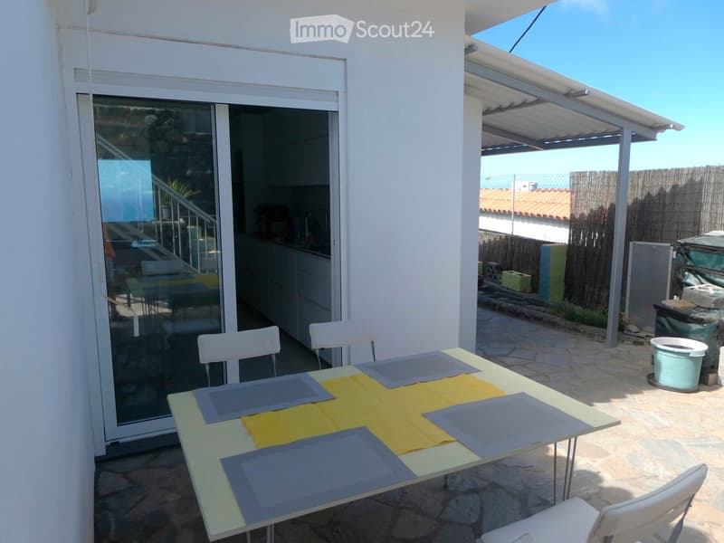 Outdoor patio with a table and chairs, covered by a pergola, adjacent to a modern house with large glass doors.