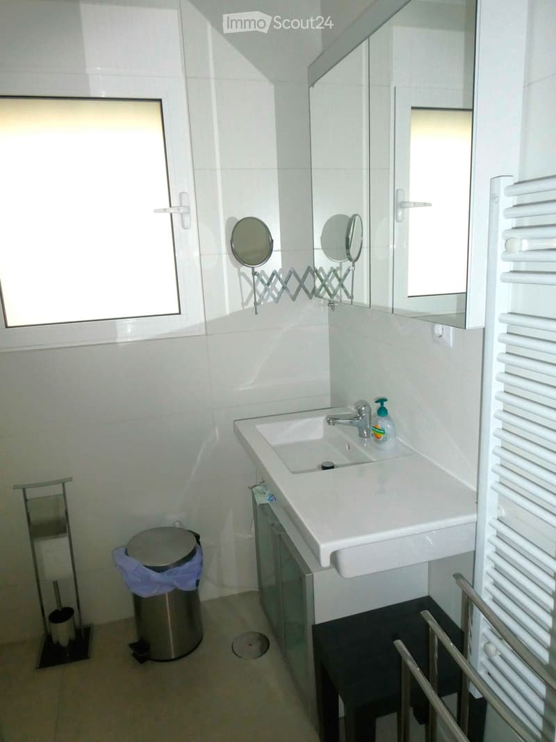 Modern bathroom with white tiles, a sink with a faucet, a mirror, a radiator, a trash can, and a small stool.