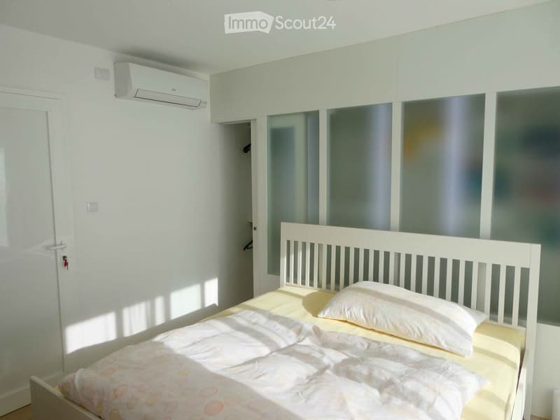 A modern, minimalist bedroom with a white bed, matching headboard, and two pillows. The room features white walls, a built-in wardrobe, and an air conditioning unit mounted on the wall.