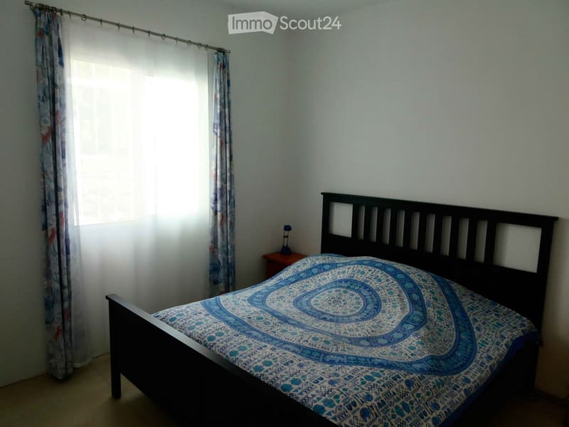 Bedroom with a dark wooden bed, blue patterned bedspread, nightstand, and lamp. Light-colored walls and curtains.