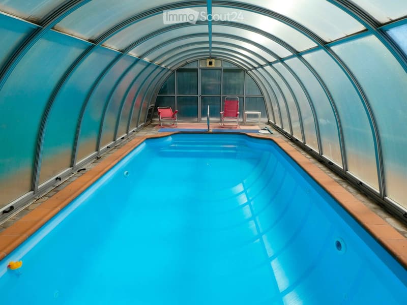 Indoor swimming pool area with blue water, surrounded by glass walls, featuring two red chairs and a small table.