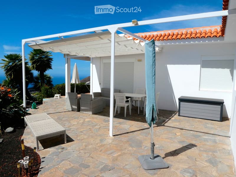 Outdoor patio with stone tiles, white pergola, sofa, table, chairs, and umbrella, overlooking the ocean.