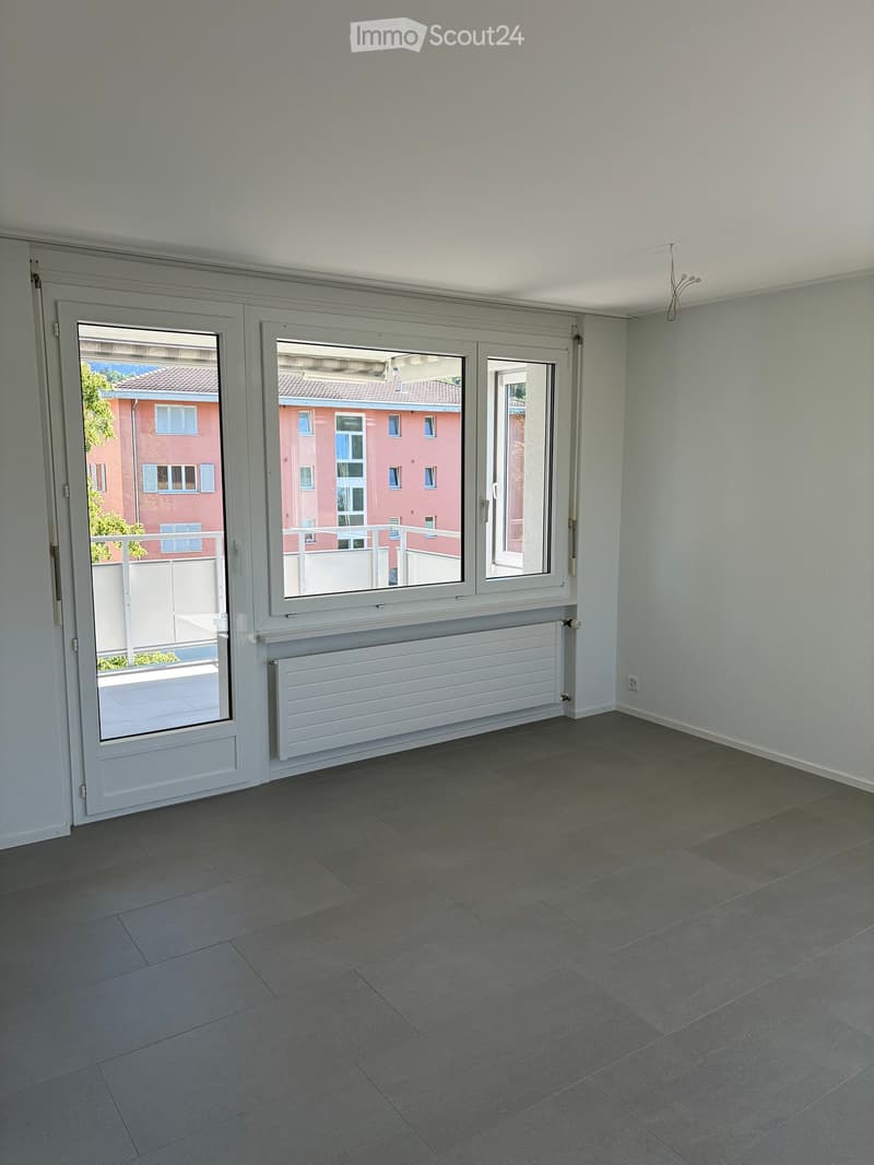 Empty room with grey tiled floor, white walls, large window with white frame, radiator, and white sliding door leading to balcony.
