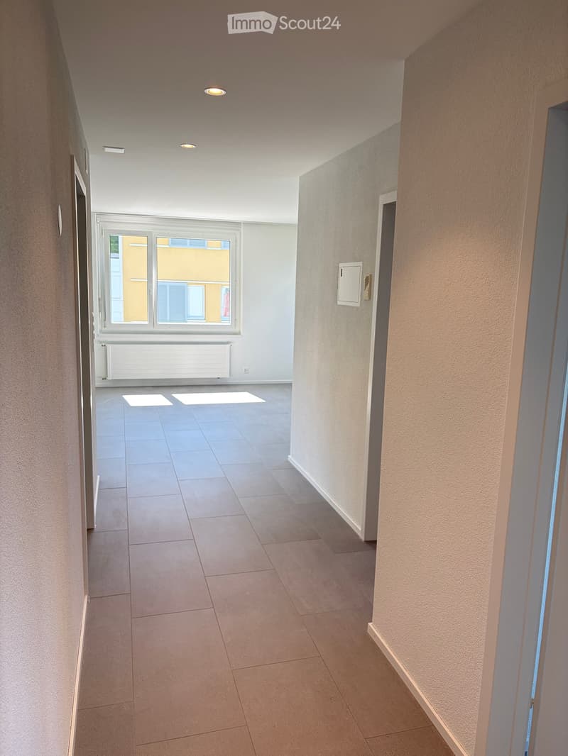 Empty hallway with tiled floor, beige walls, and a window at the end showing a view of a building.