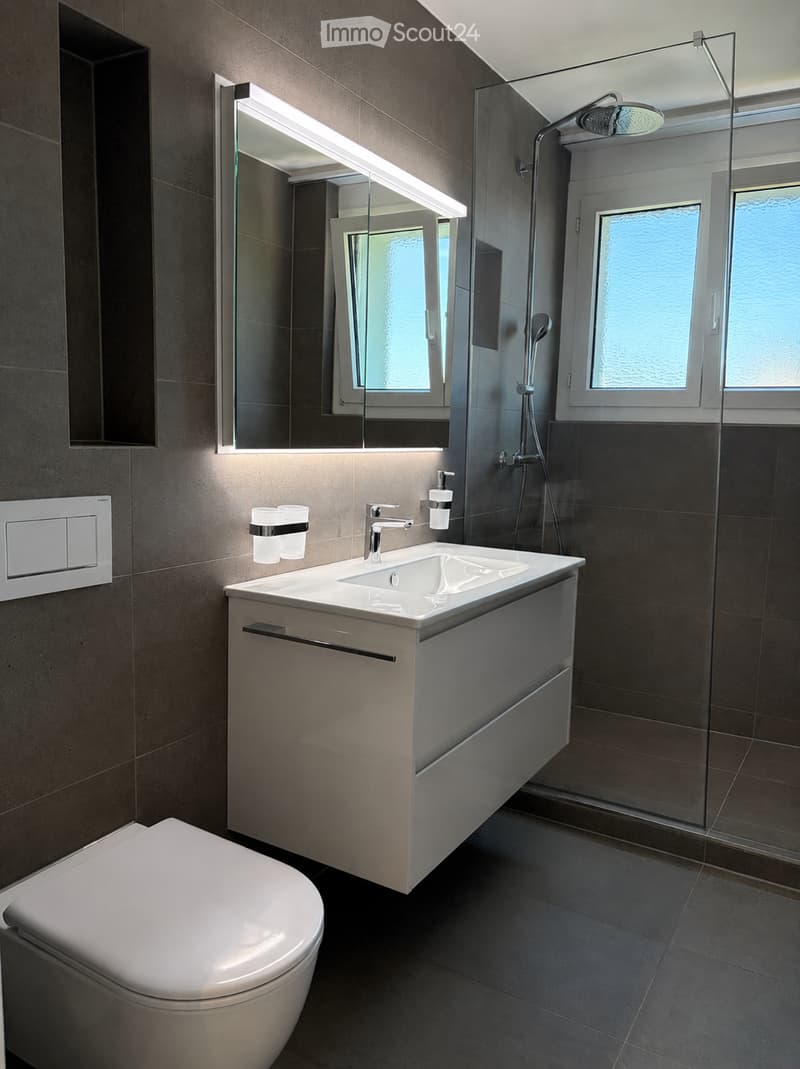 Modern bathroom with a white sink, chrome faucet, toilet, large mirror with built-in lighting, and a glass-enclosed shower area.