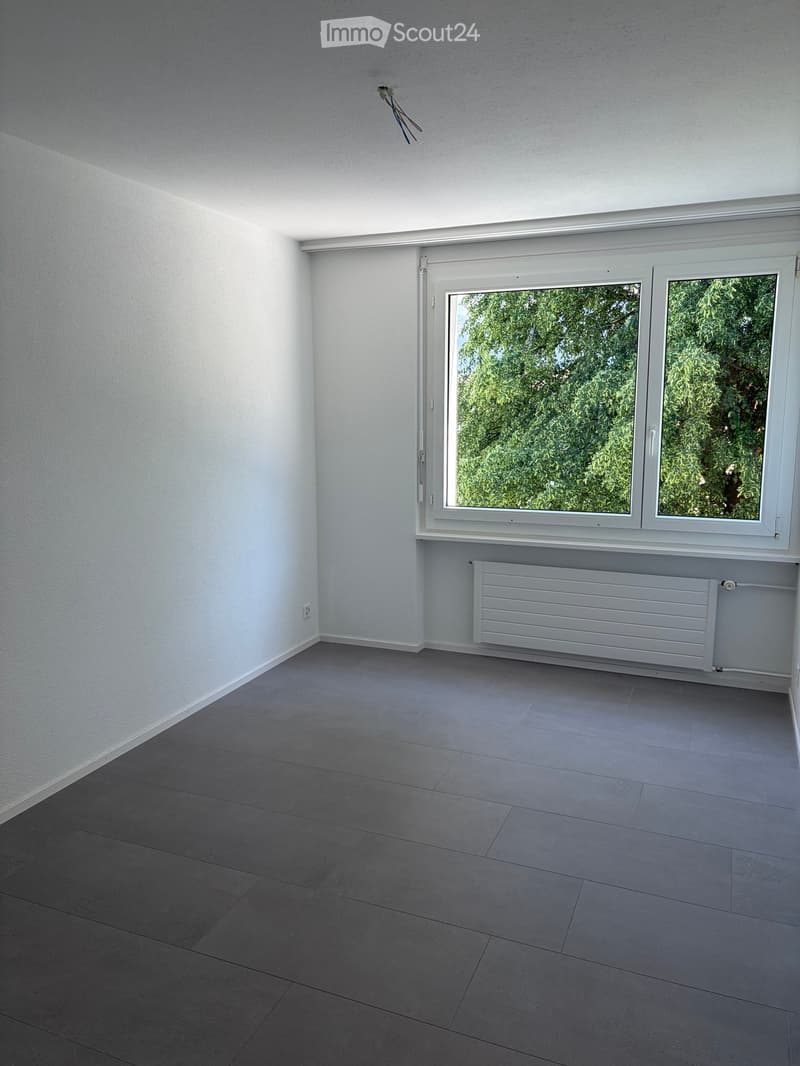 Empty room with light grey tiled floor, white walls, large window with white frame, radiator below window, and electrical outlet on wall.