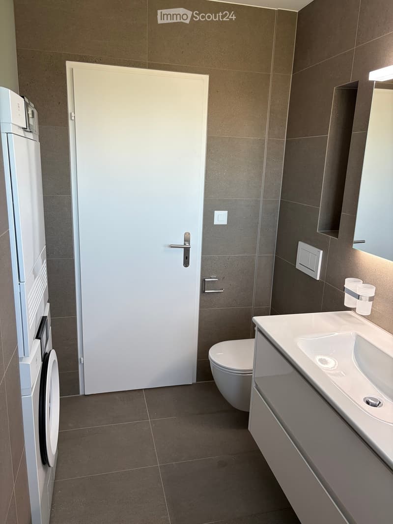 Modern bathroom with a white sink, toilet, and two cups on the counter. The room features tiled walls and a door leading to another area.