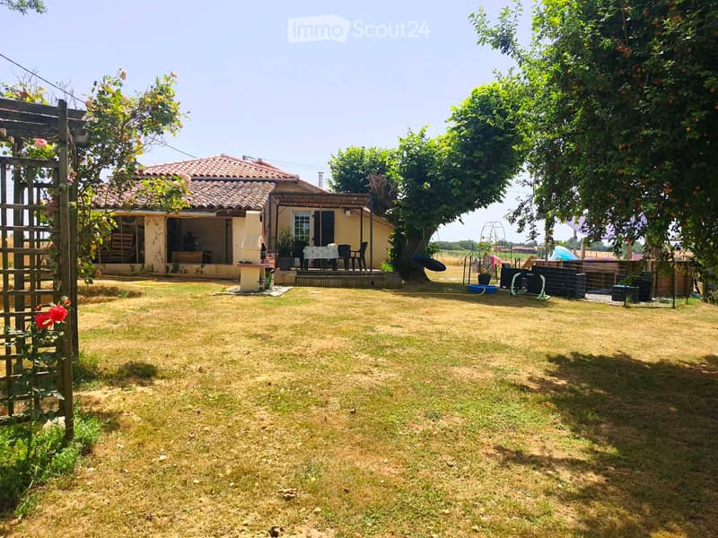 A sunny garden area with a small house, patio furniture, and trees providing shade.